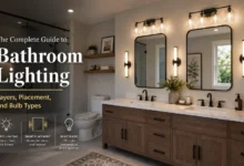 Modern bathroom featuring layered lighting with a halo-lit round mirror, wall sconces, recessed ceiling lights, a glass walk-in shower, freestanding bathtub, and an infographic explaining ambient, task, and accent lighting alongside a color temperature guide for bathroom lighting.